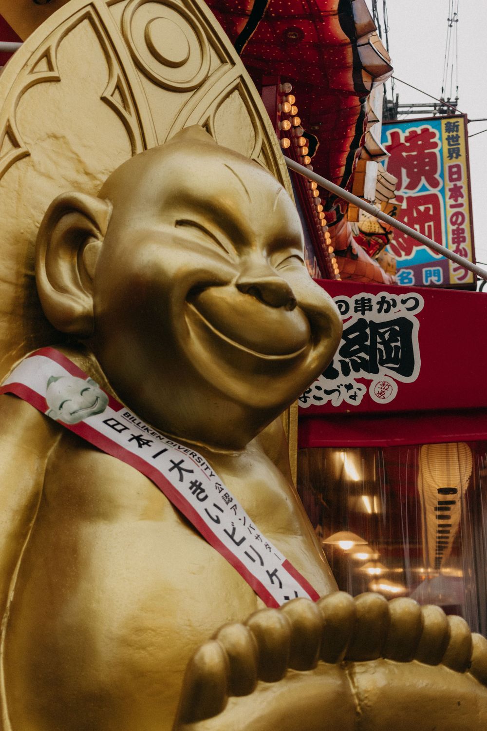 Golden lucky Buddha statue with a smiling face against Japanese signage and decorations.