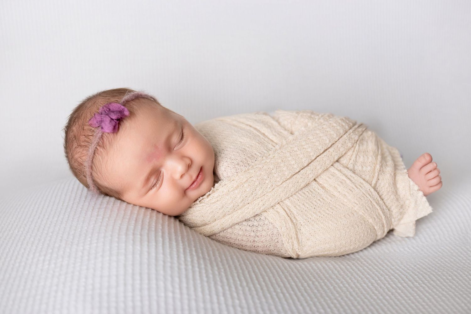 Peaceful sleeping newborn wrapped in cream-colored swaddle on soft gray blanket with purple floral headband.