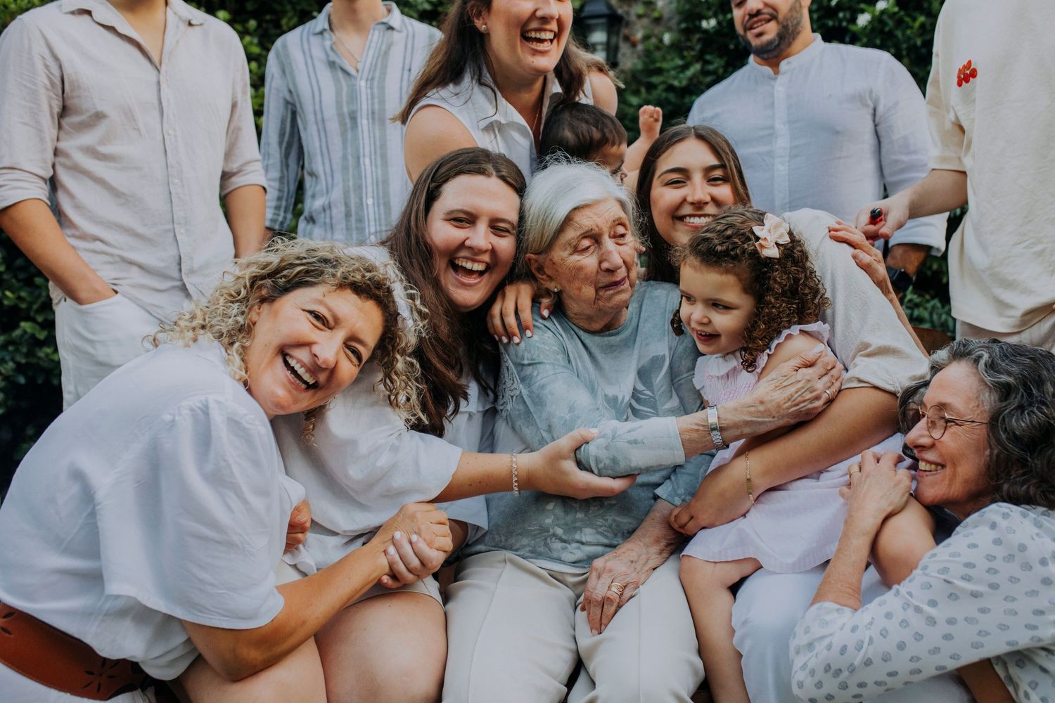A joyful multi-generational family laughs together while sitting outdoors in casual light-colored clothing.
