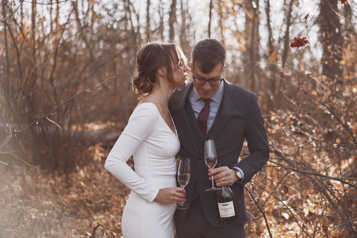 A newlywed couple shares a joyful, intimate moment in an autumn forest, toasting with champagne as golden sunlight filters through the trees