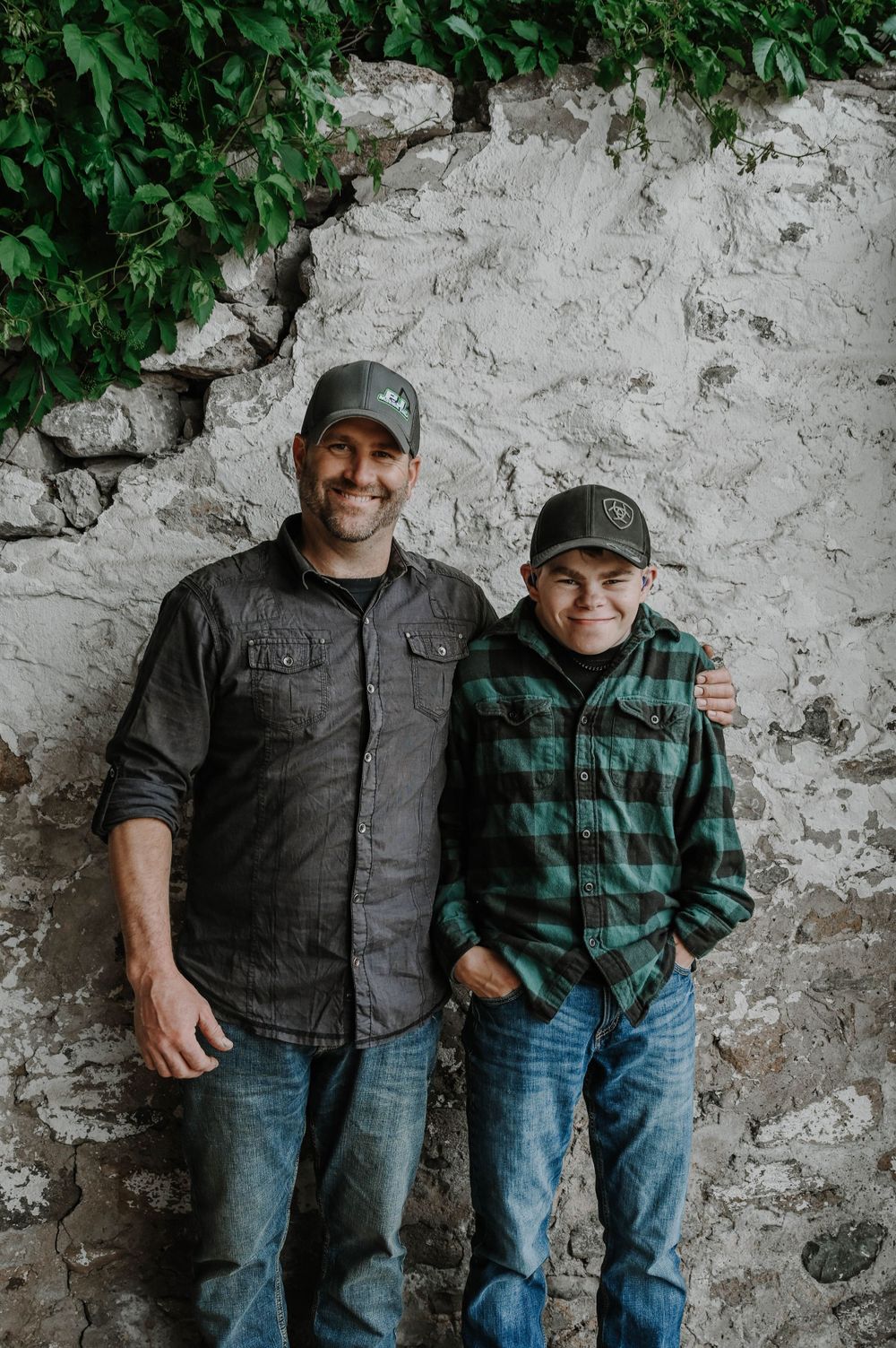 Two people in baseball caps and casual clothing pose against a white stone wall with green foliage visible.