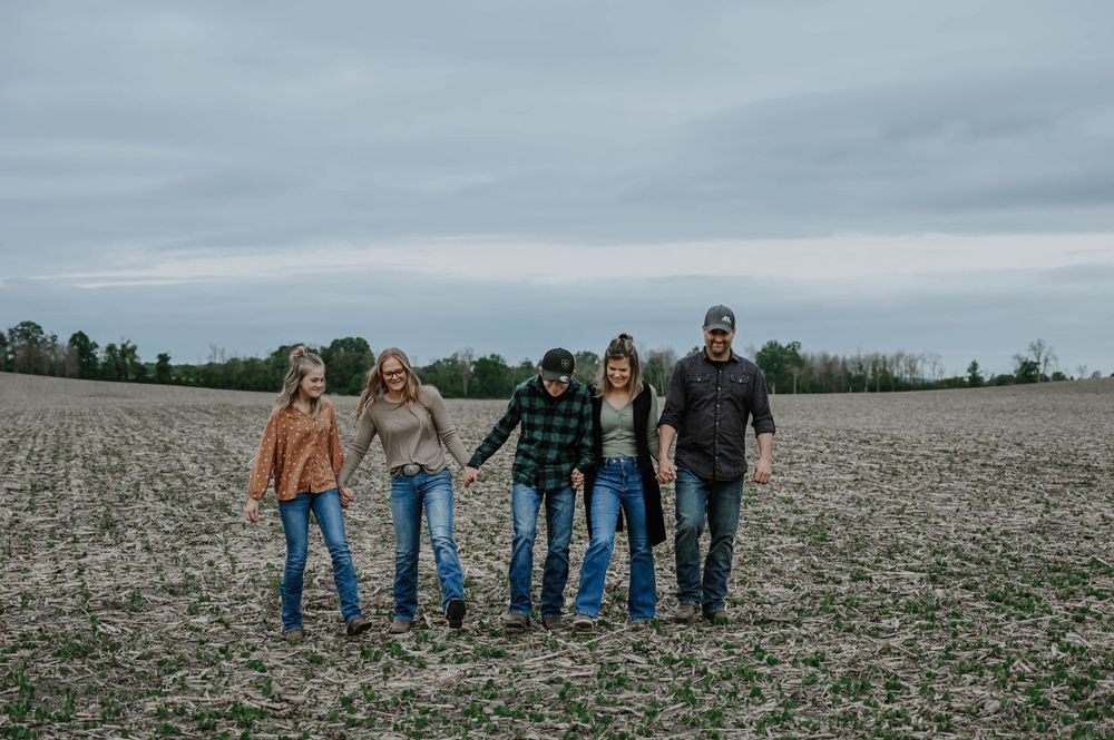 A group of people stand together in a plowed field on an overcast day with trees in the background.