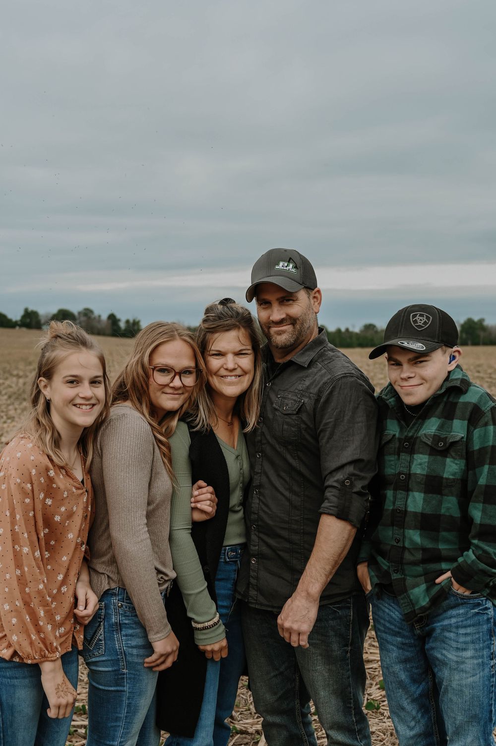 A group of five people smiling and posing together on a rural field against an overcast sky.
