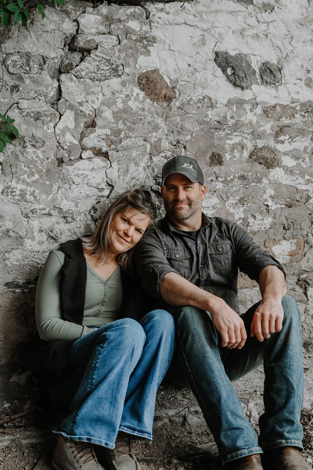 A couple sits casually against a weathered stone wall background in casual attire and denim jeans.