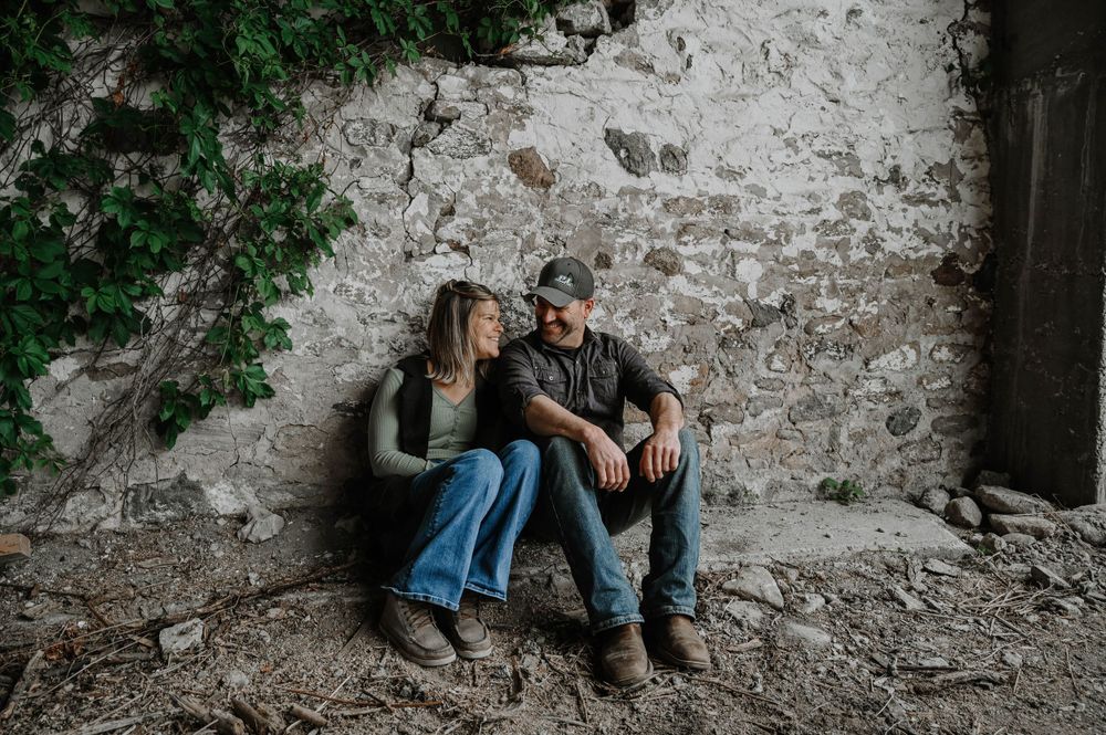 Two people sitting together against a weathered stone wall with green foliage peeking in from above.