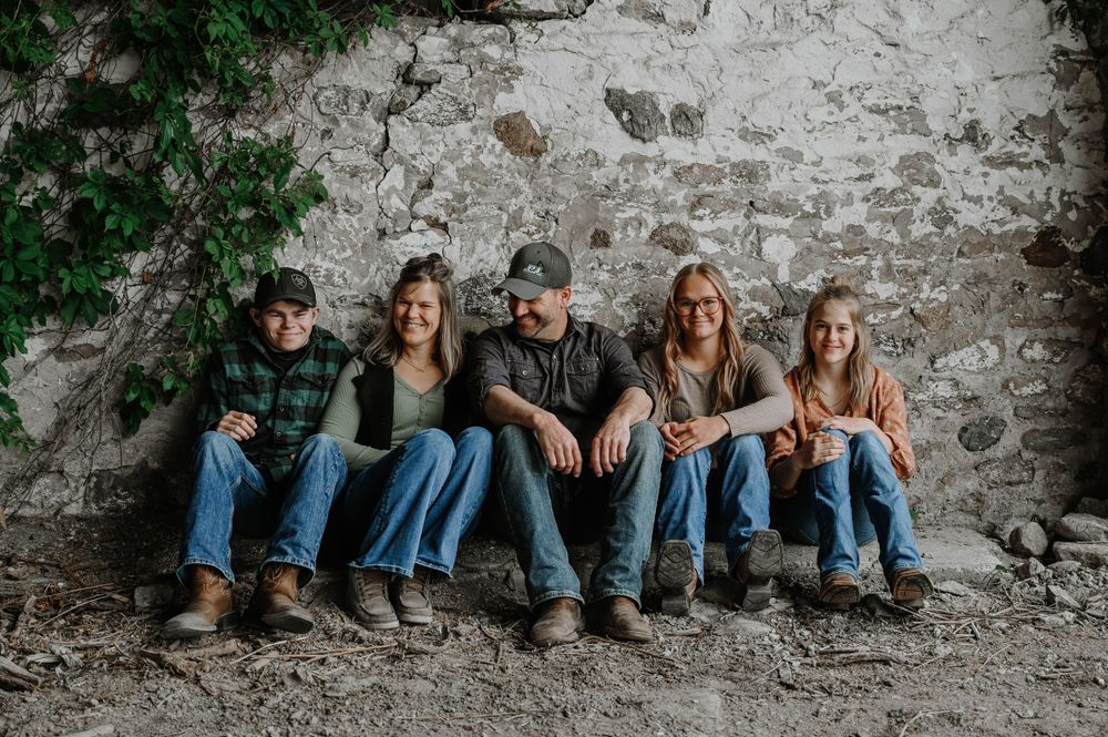 Five people in casual denim attire sitting together against a rustic stone wall with green foliage.
