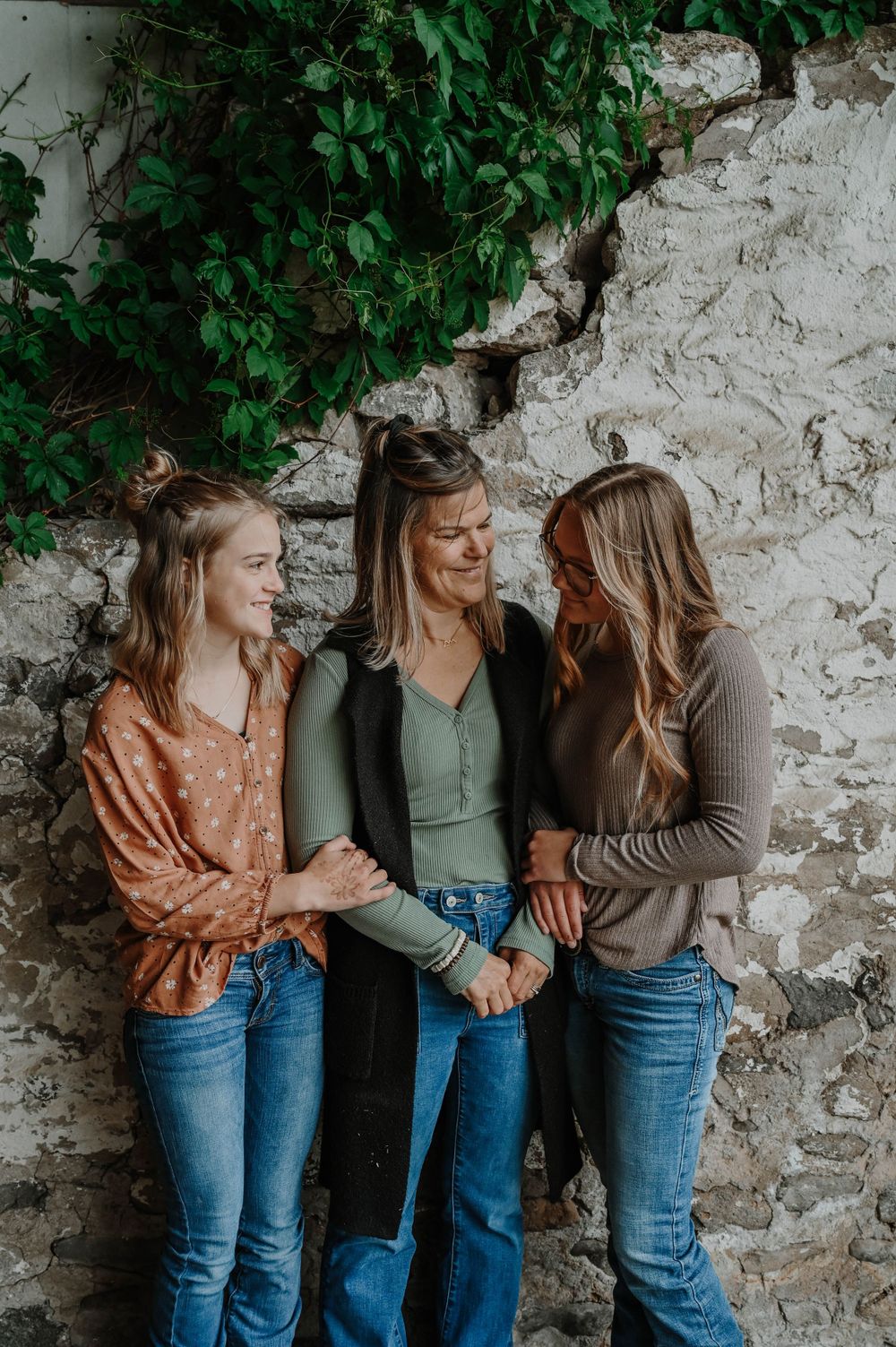 Three friends in casual fall outfits lean against a stone wall with green vines while sharing a candid moment together.