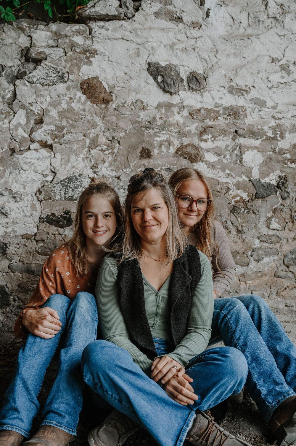 Three people in casual denim and sweaters sit together against a weathered stone wall in a natural portrait setting.