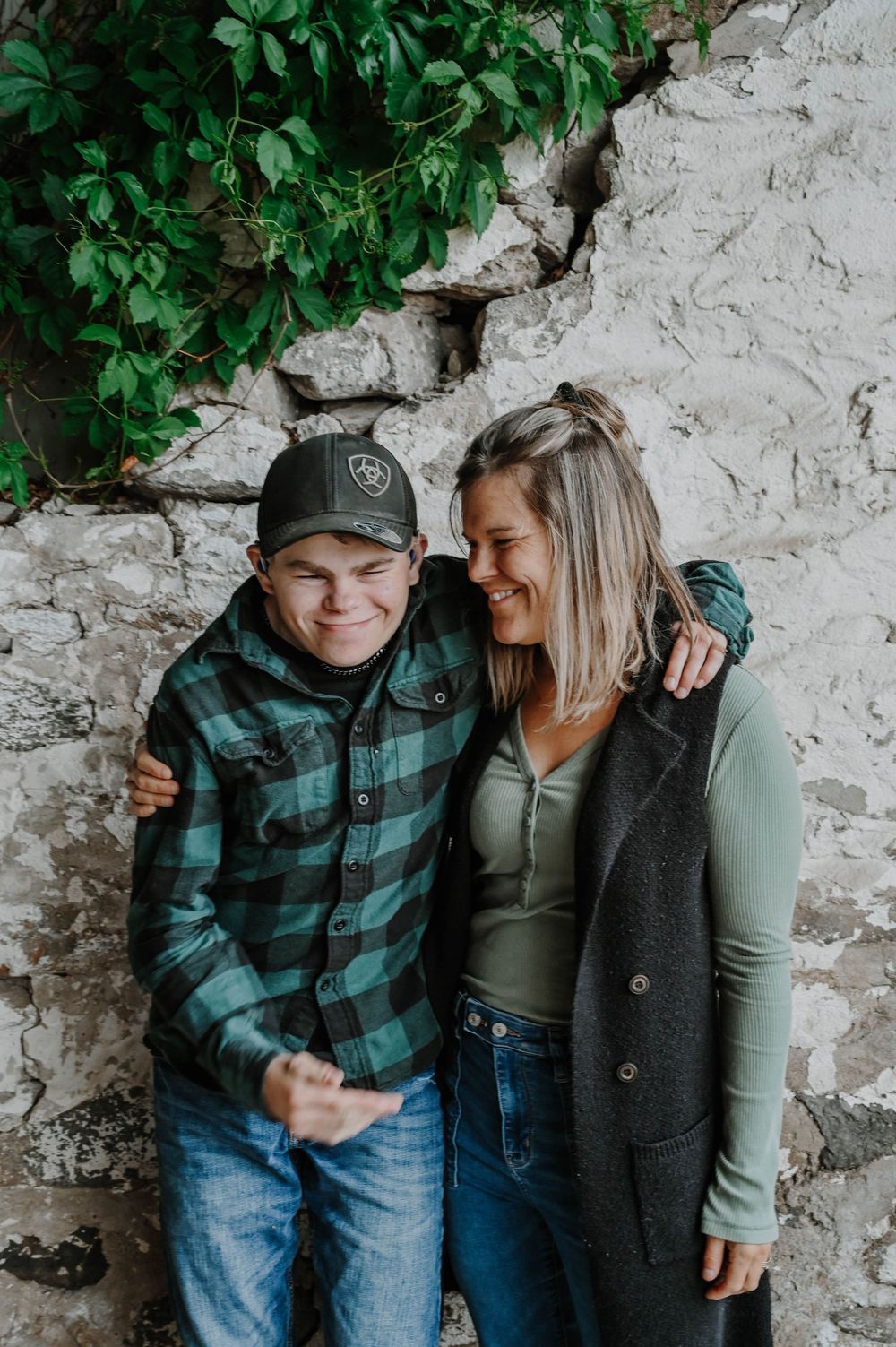 A couple laughs together against a stone wall, wearing casual fall clothing including a plaid flannel and cozy sweater.