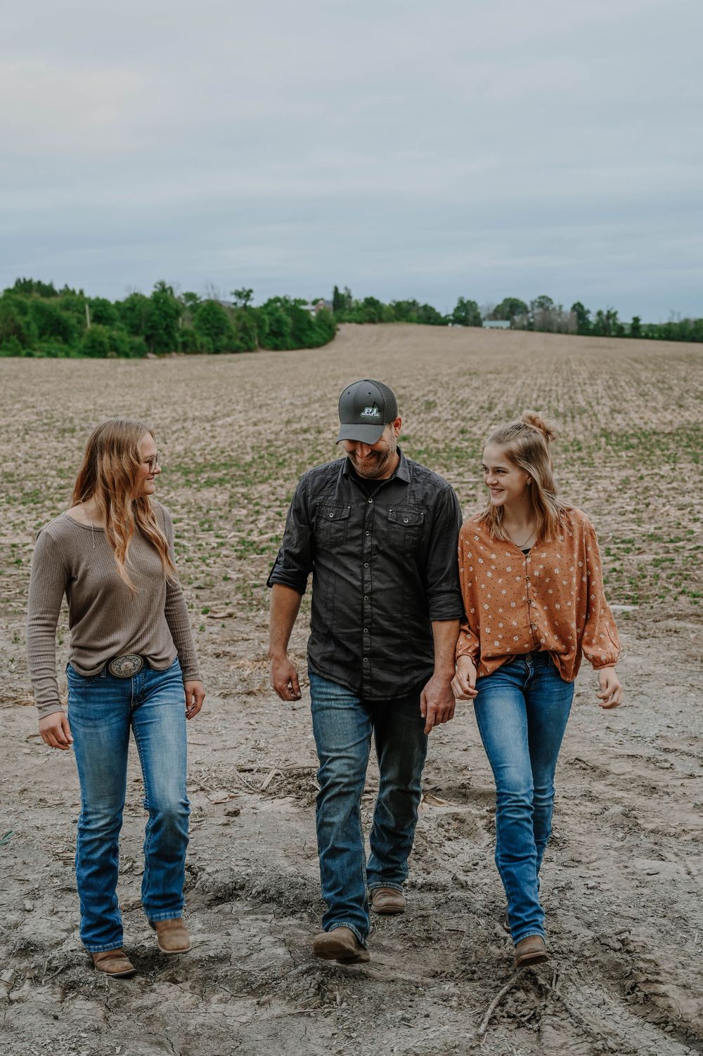 Three people walk together on a plowed dirt field with trees and cloudy sky in the background.
