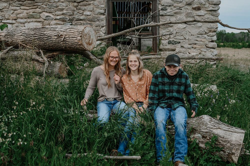 Three people sit together on a fallen log outside a rustic stone building surrounded by wildflowers.