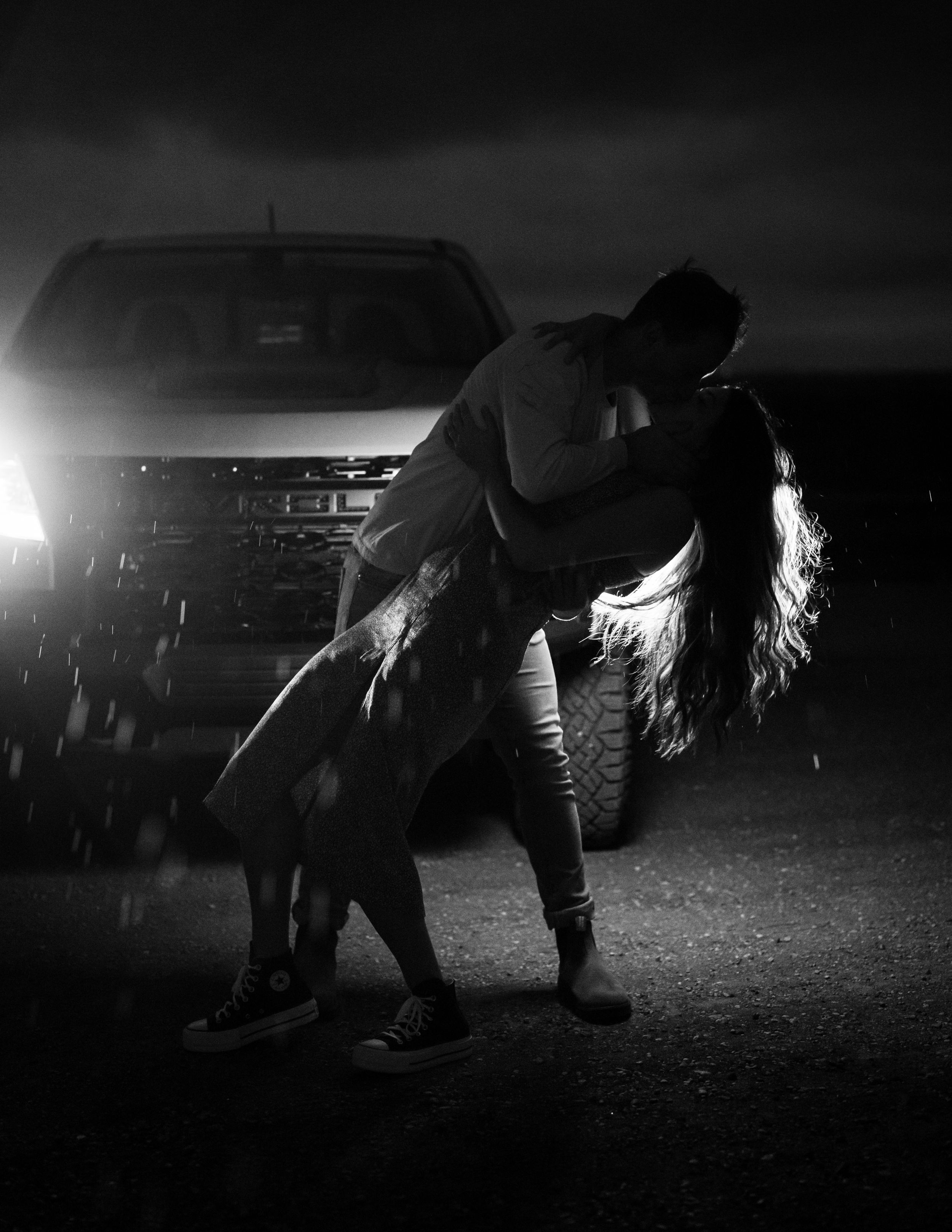 A dramatic black and white silhouette of a romantic dip pose in front of car headlights in the rain.