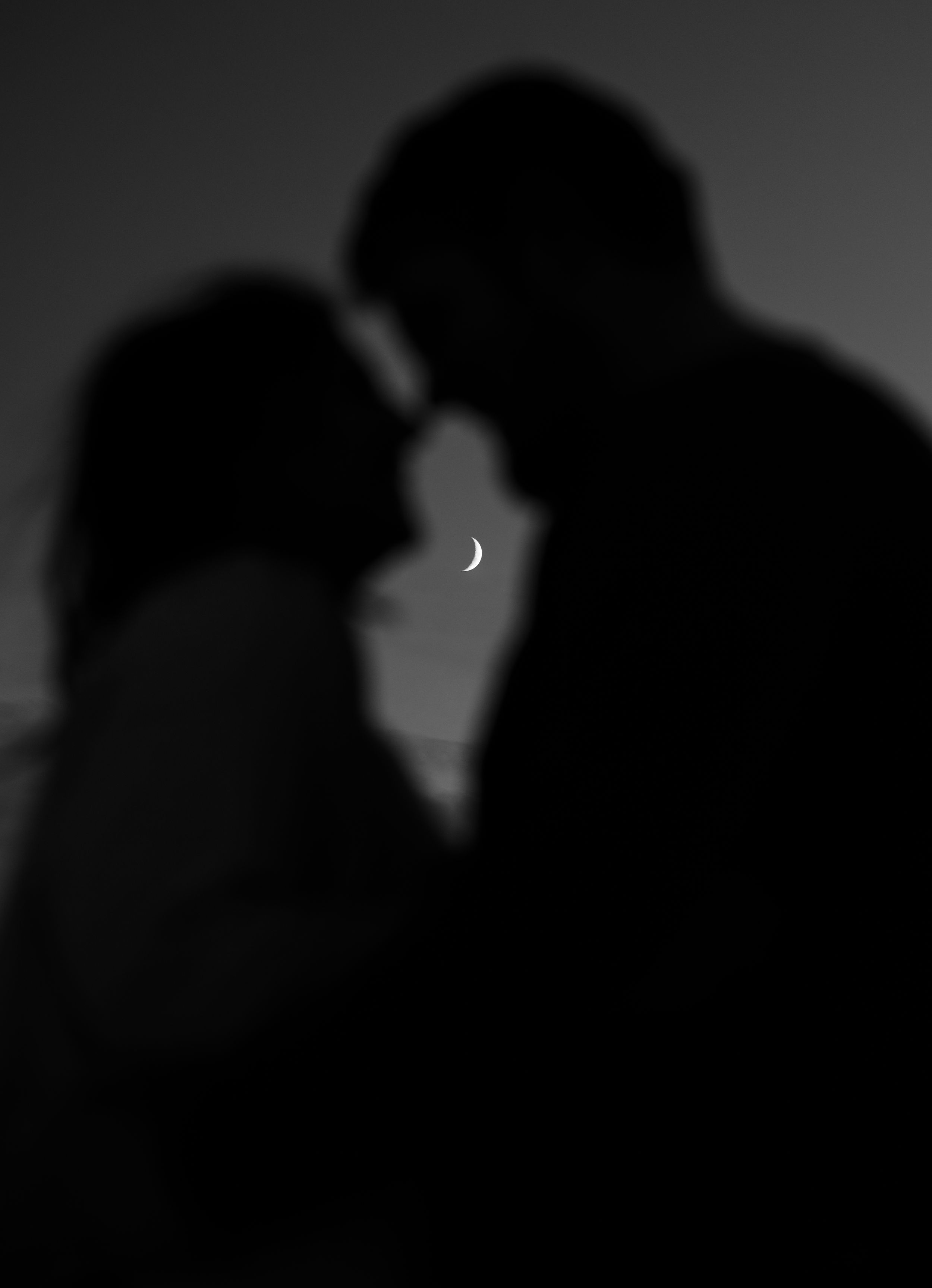 Silhouettes of a couple about to kiss with a crescent moon visible between them against a dark background.