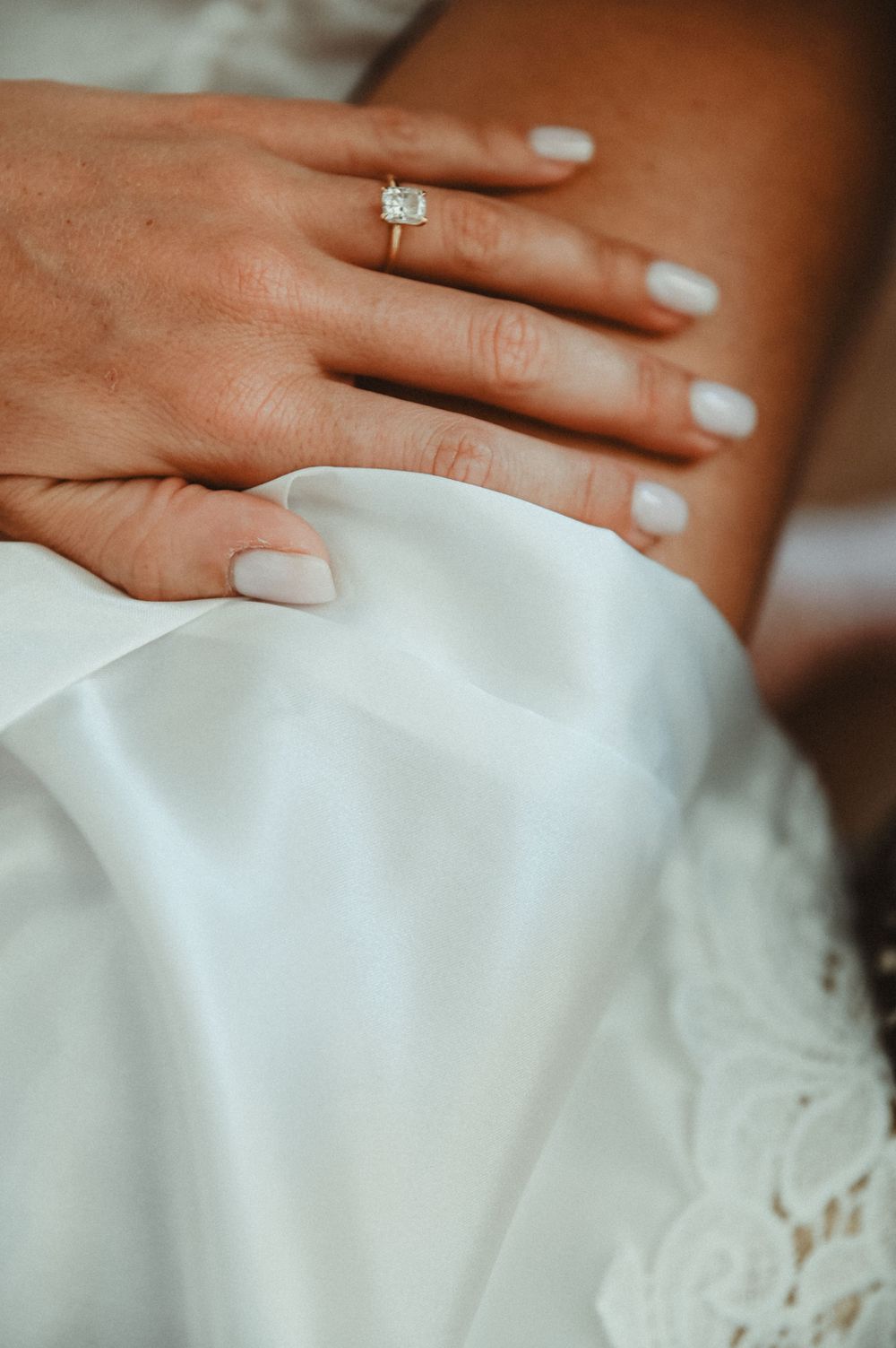 A hand with light nail polish and a sparkling engagement ring rests on white satin and lace fabric.