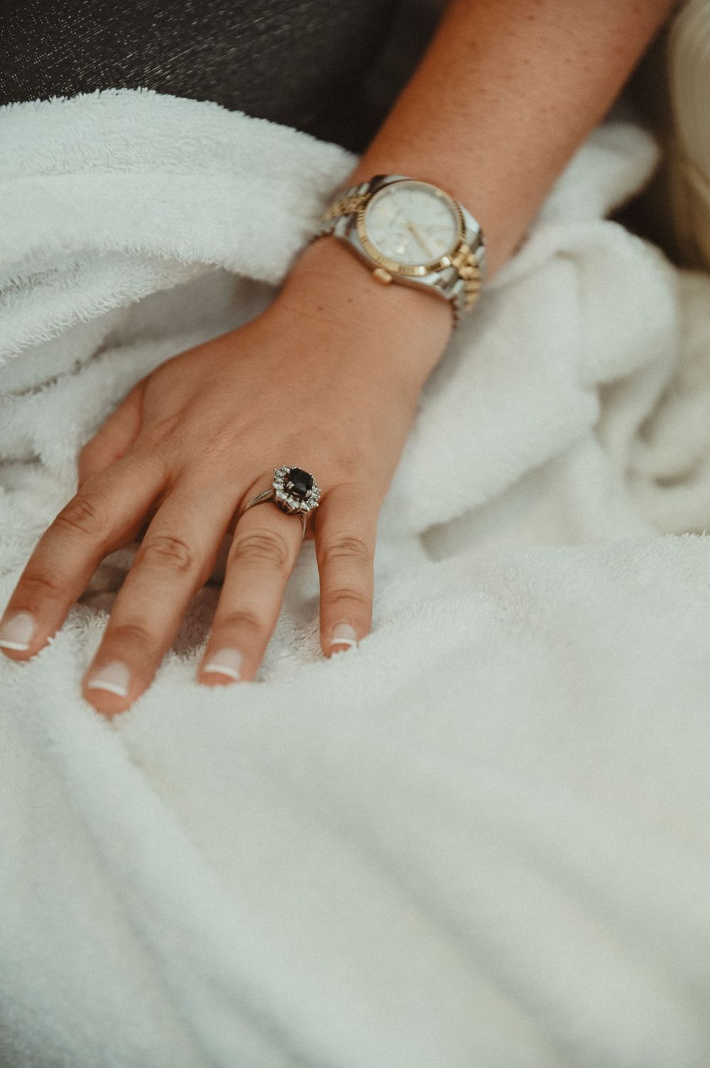 Close-up of a hand wearing a vintage watch and dark gemstone ring against white fabric.