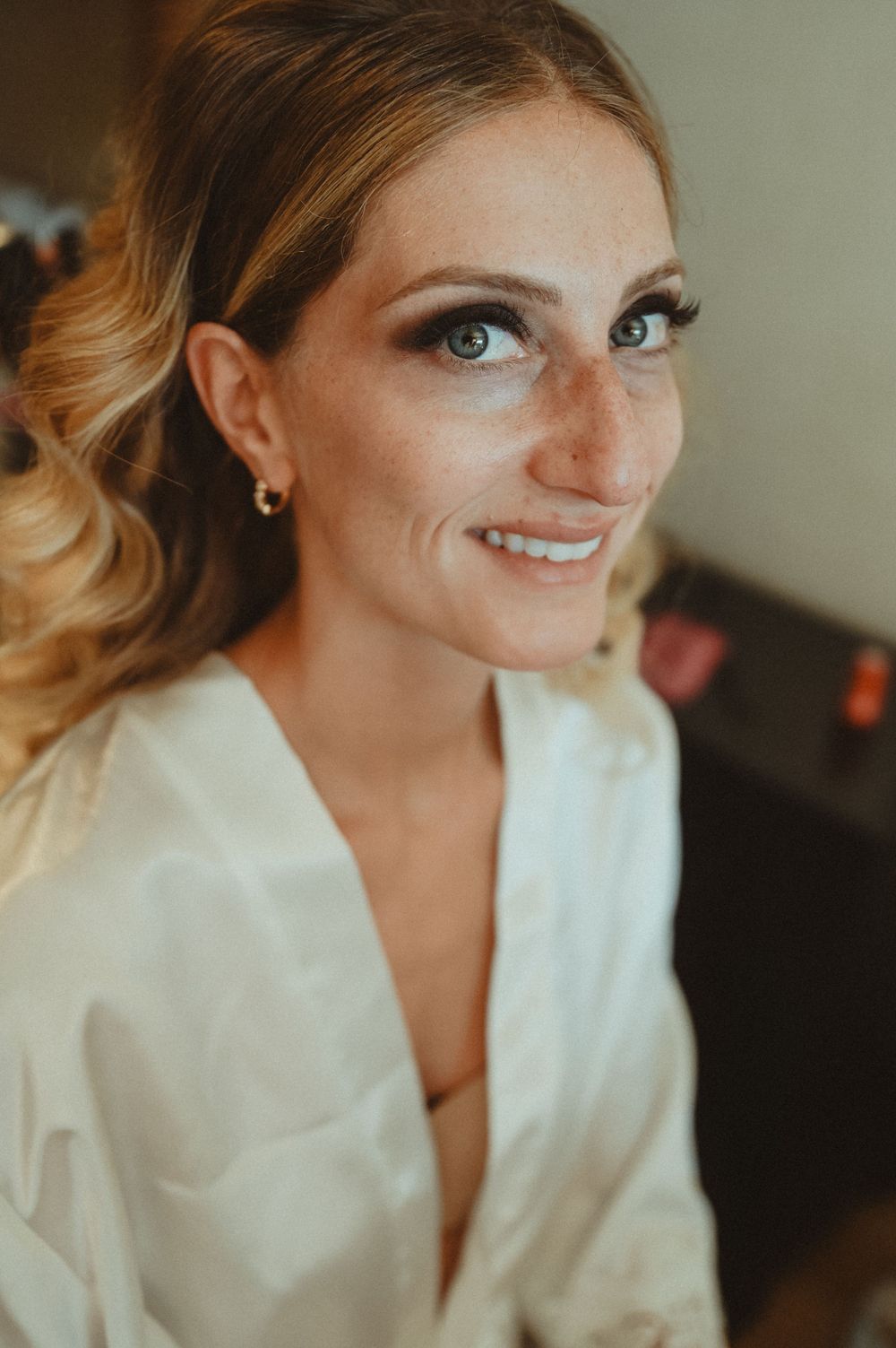 Close-up portrait of someone with blonde hair and bright blue eyes wearing a white robe and smiling warmly at the camera.