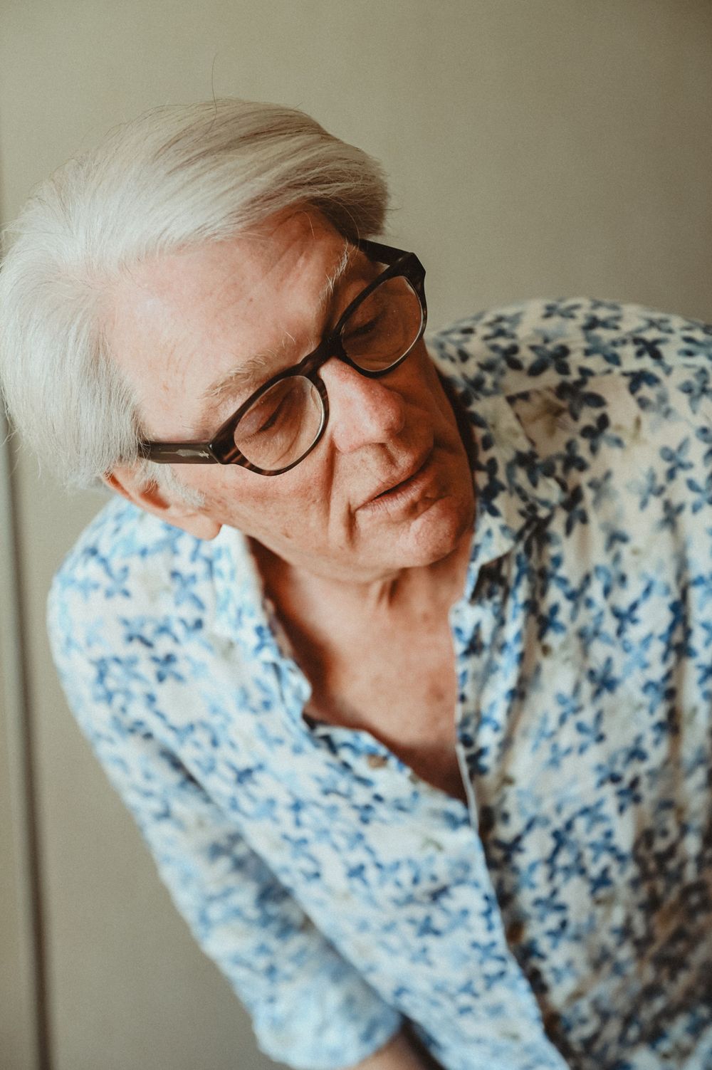 An elderly person wearing glasses and a blue floral patterned shirt smiles warmly in a natural light setting.