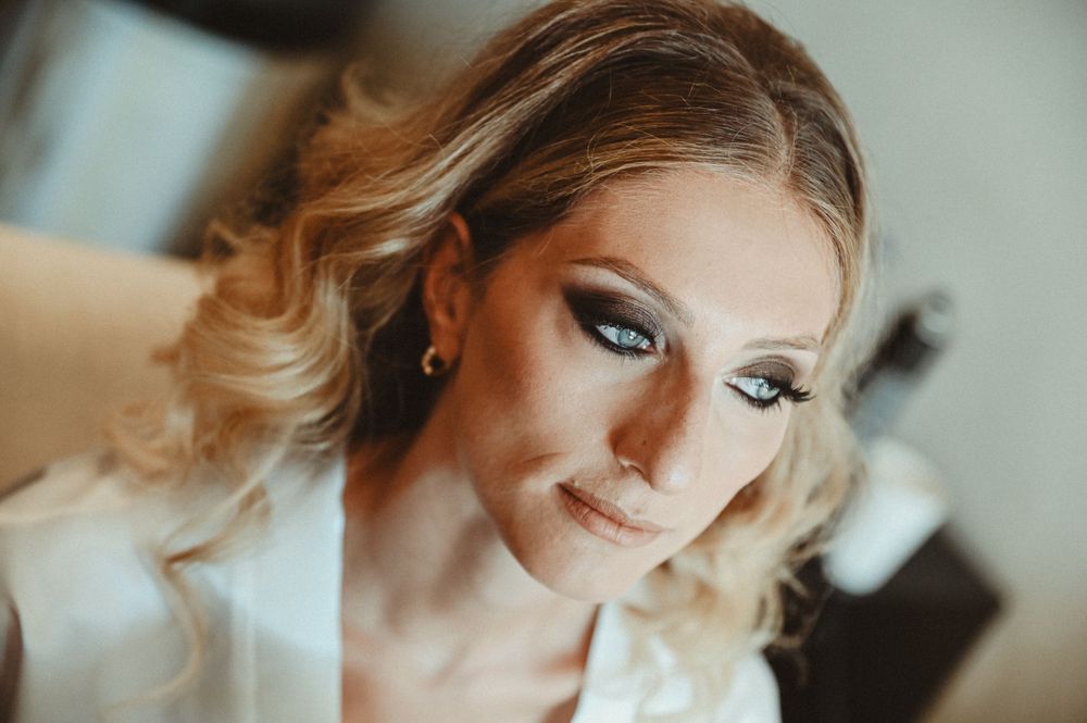 A close-up beauty portrait showing glamorous makeup, blonde wavy hair, and sparkly earrings in soft lighting.