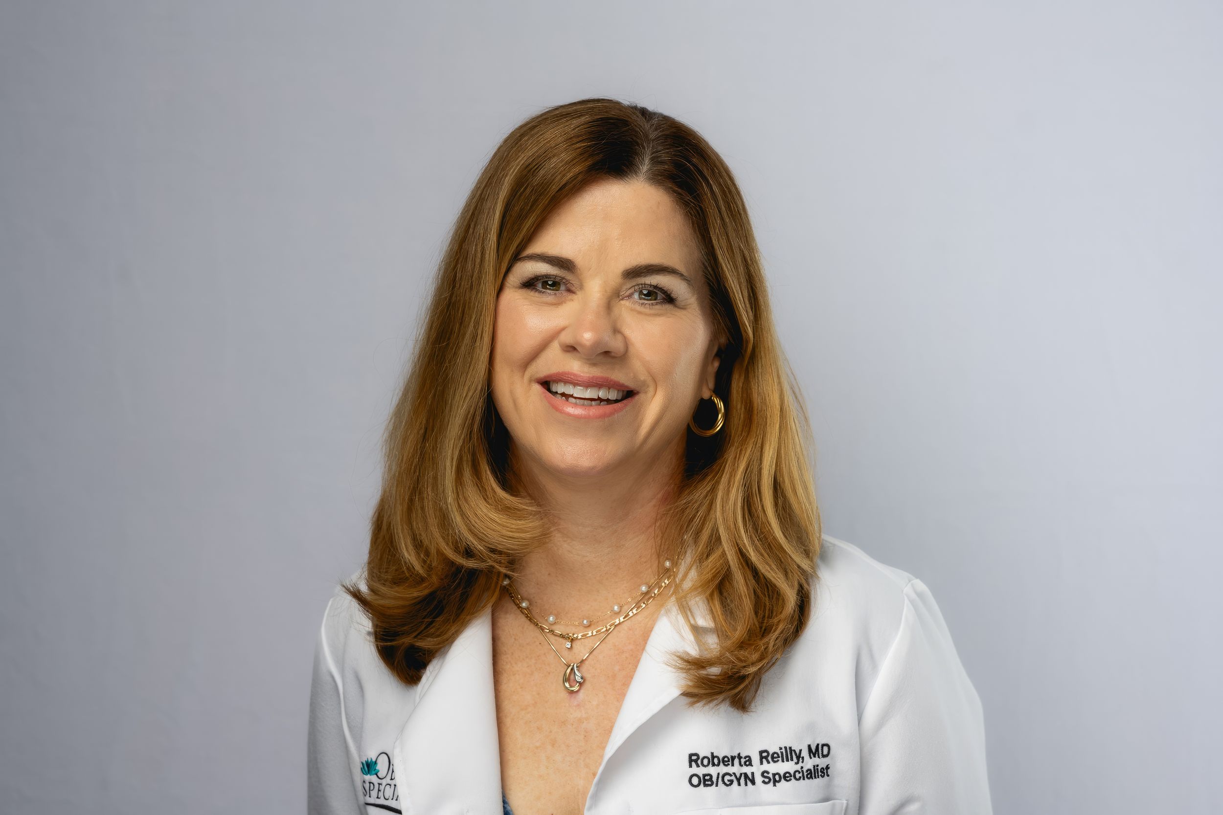 Professional healthcare provider in white medical coat smiling against gray background.