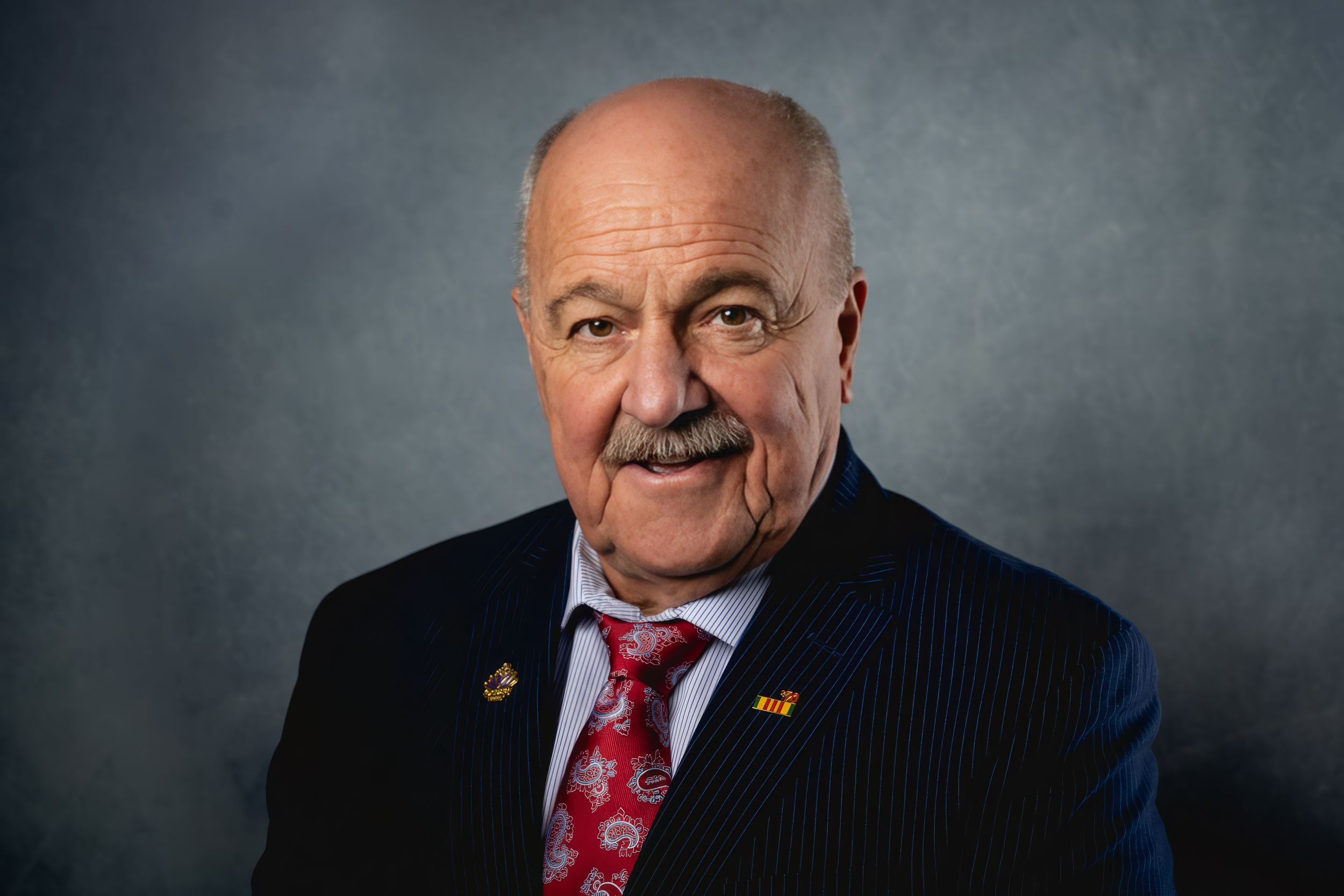 Professional portrait of someone in a dark pinstripe suit with red tie against gray background.
