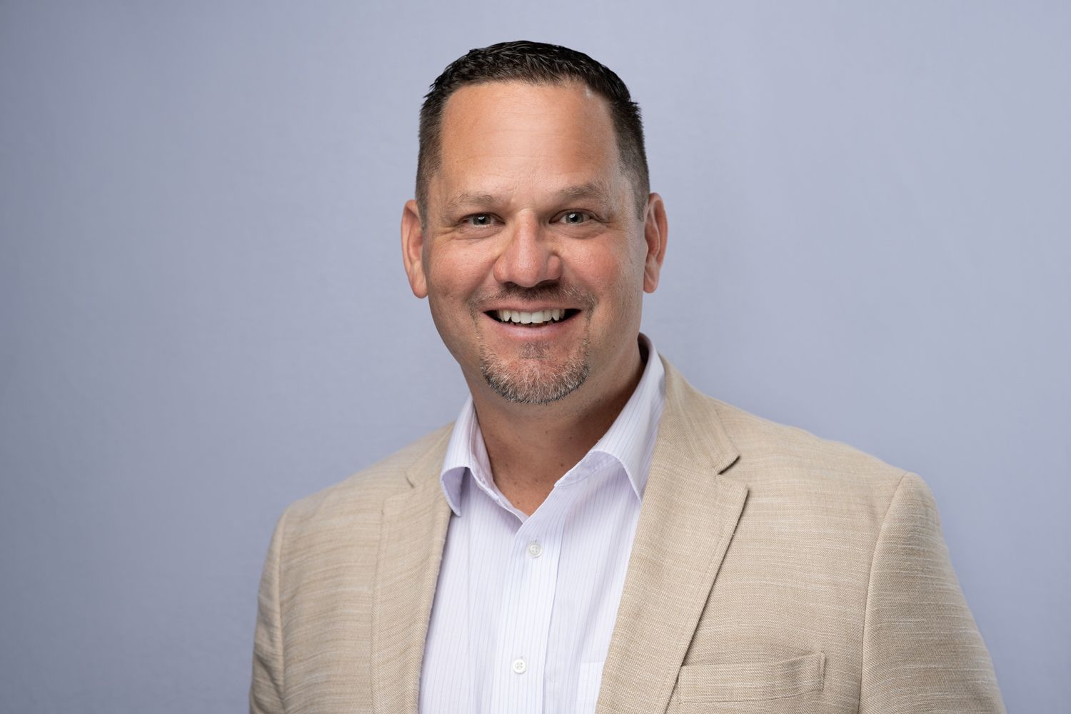 Professional headshot of a smiling man wearing a beige blazer and white dress shirt against a gray background.