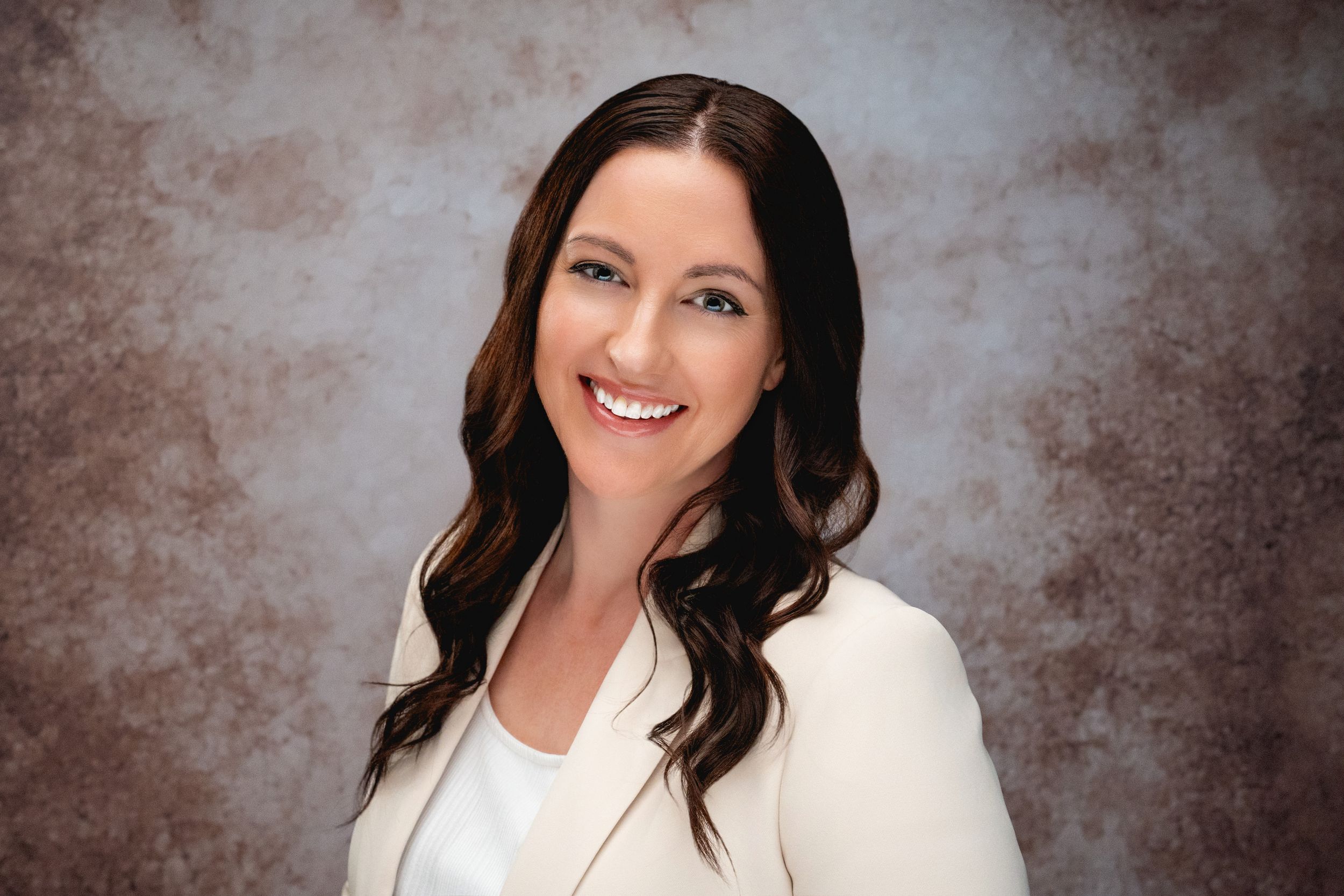 Professional headshot of a person wearing a white blazer and styling long dark hair against a brown textured backdrop.