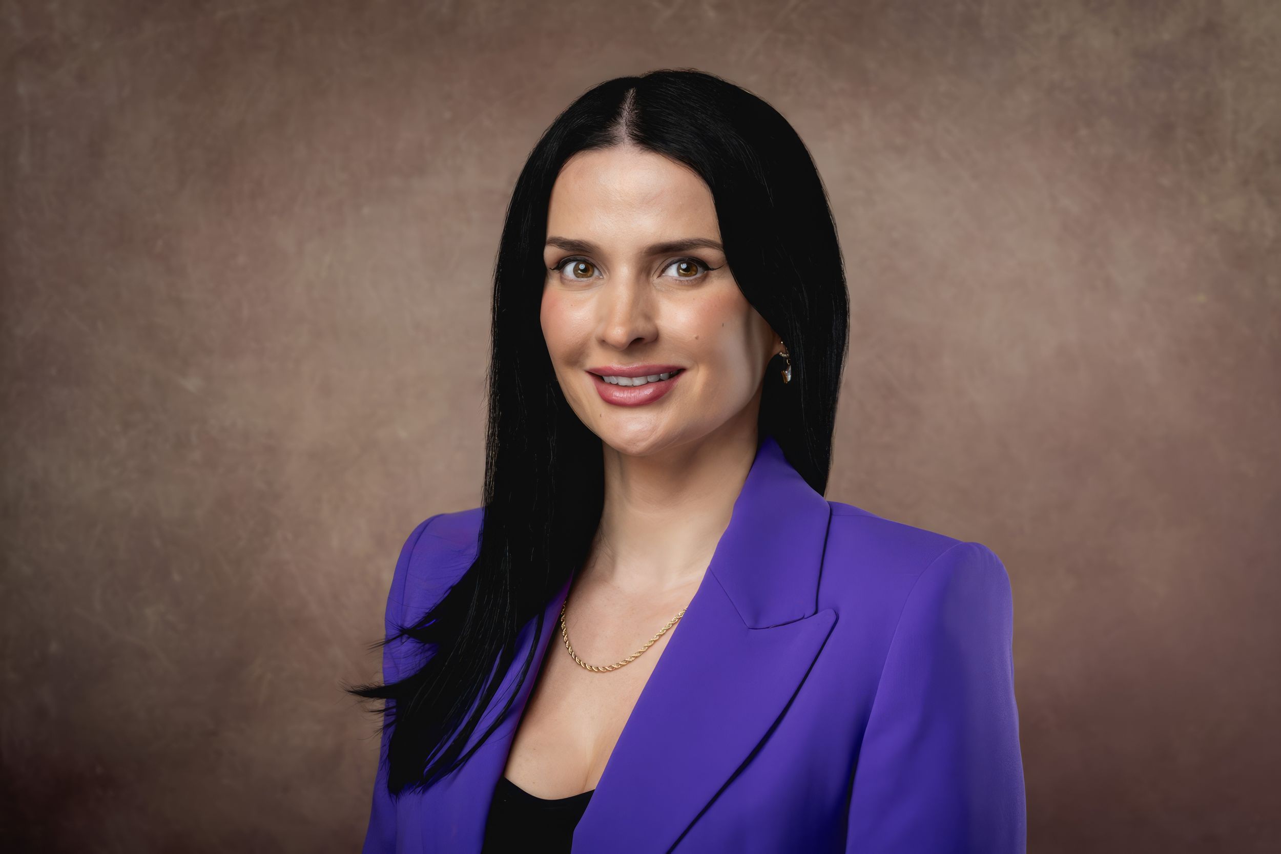 Professional portrait of someone in a vibrant purple blazer and dark hair against a textured brown backdrop.