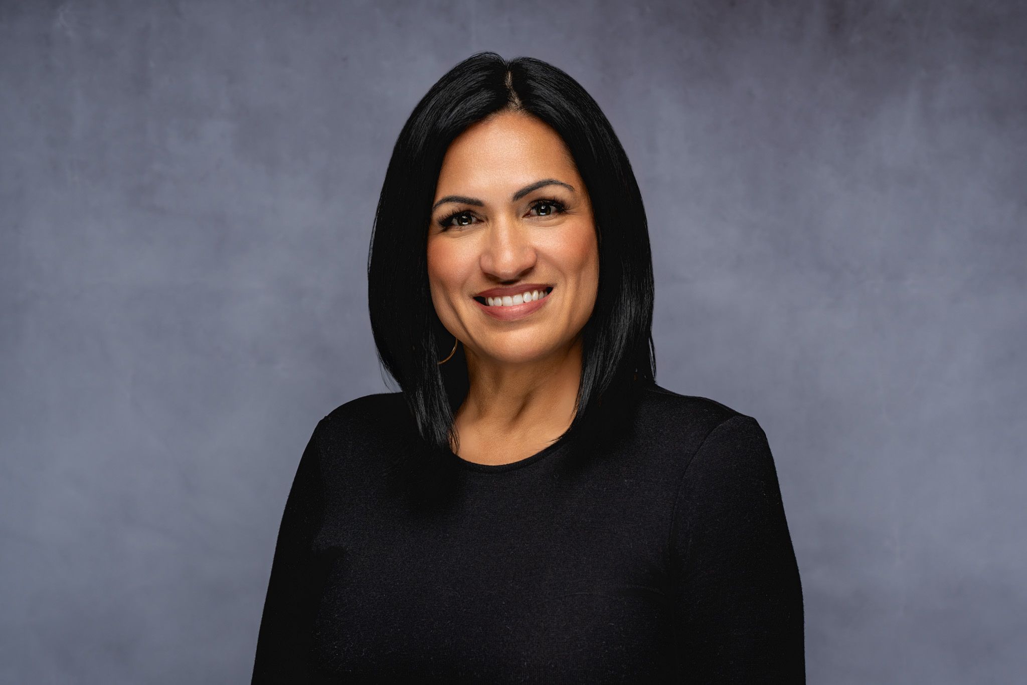 Professional headshot portrait of someone in a black top smiling warmly against a gray background.