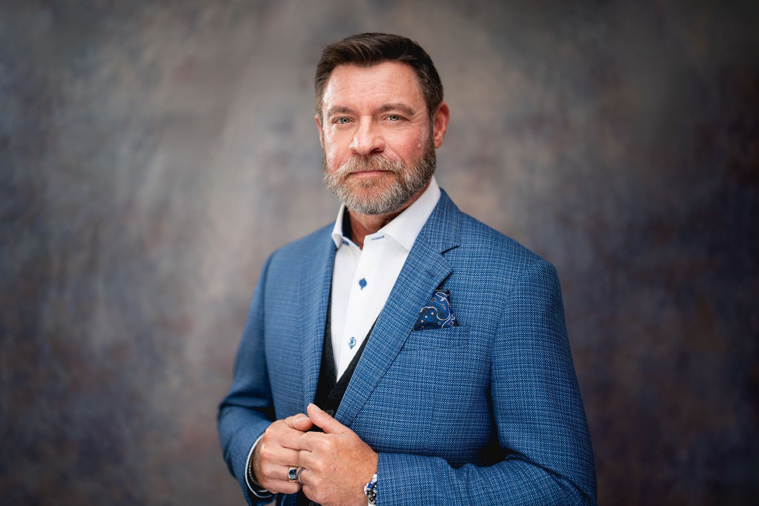 A person in a blue textured suit jacket and white shirt poses confidently against a dark studio backdrop.