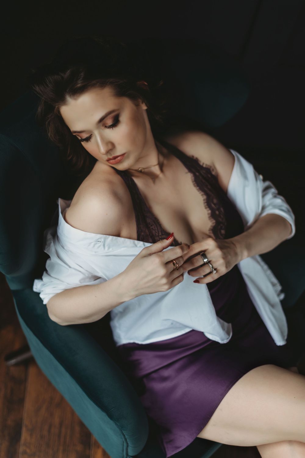 Artistic boudoir portrait in teal armchair showing figure in white top and purple skirt with moody lighting  in Bellevue studio.