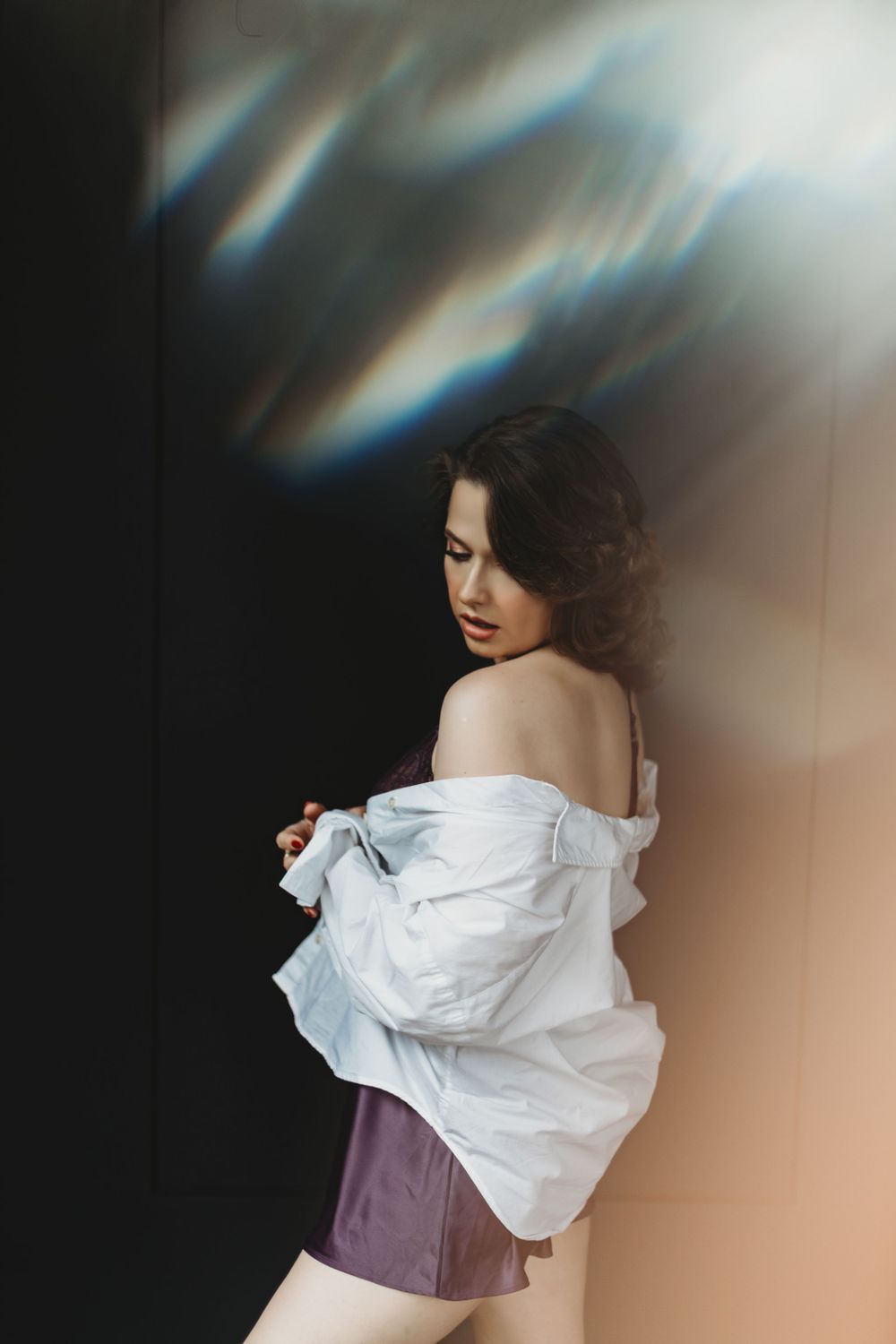 Artistic boudoir portrait with dramatic lighting shows a figure in a white shirt and purple corset against dark backdrop  in Bellevue studi.