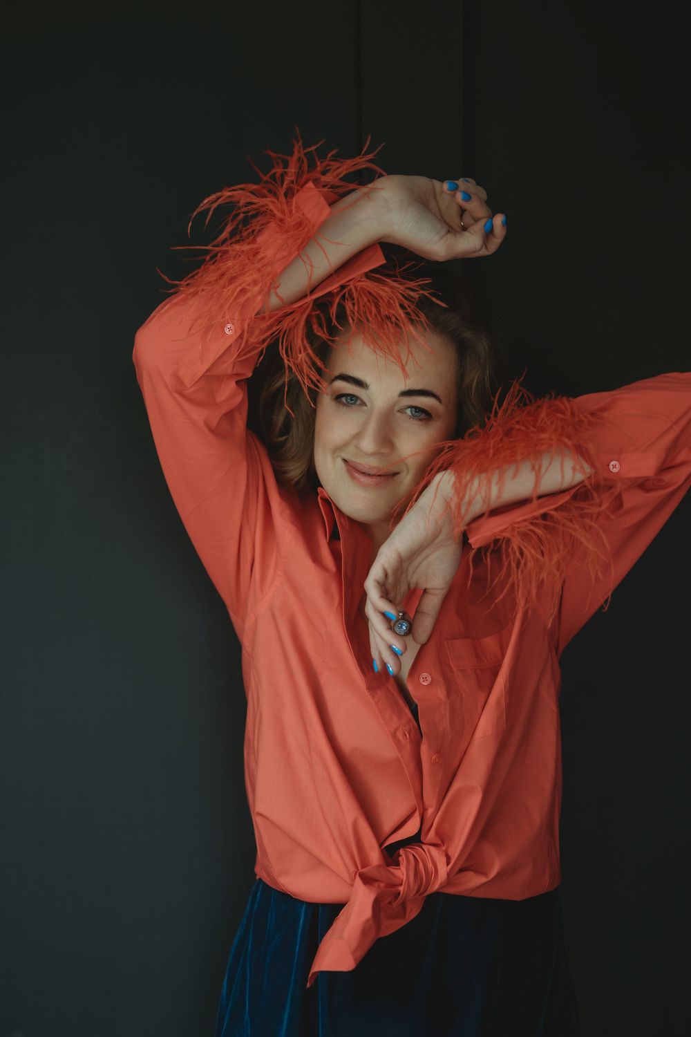 Playful fashion portrait against dark background featuring coral-colored blouse with feather details and blue skirt in Bellevue studio.