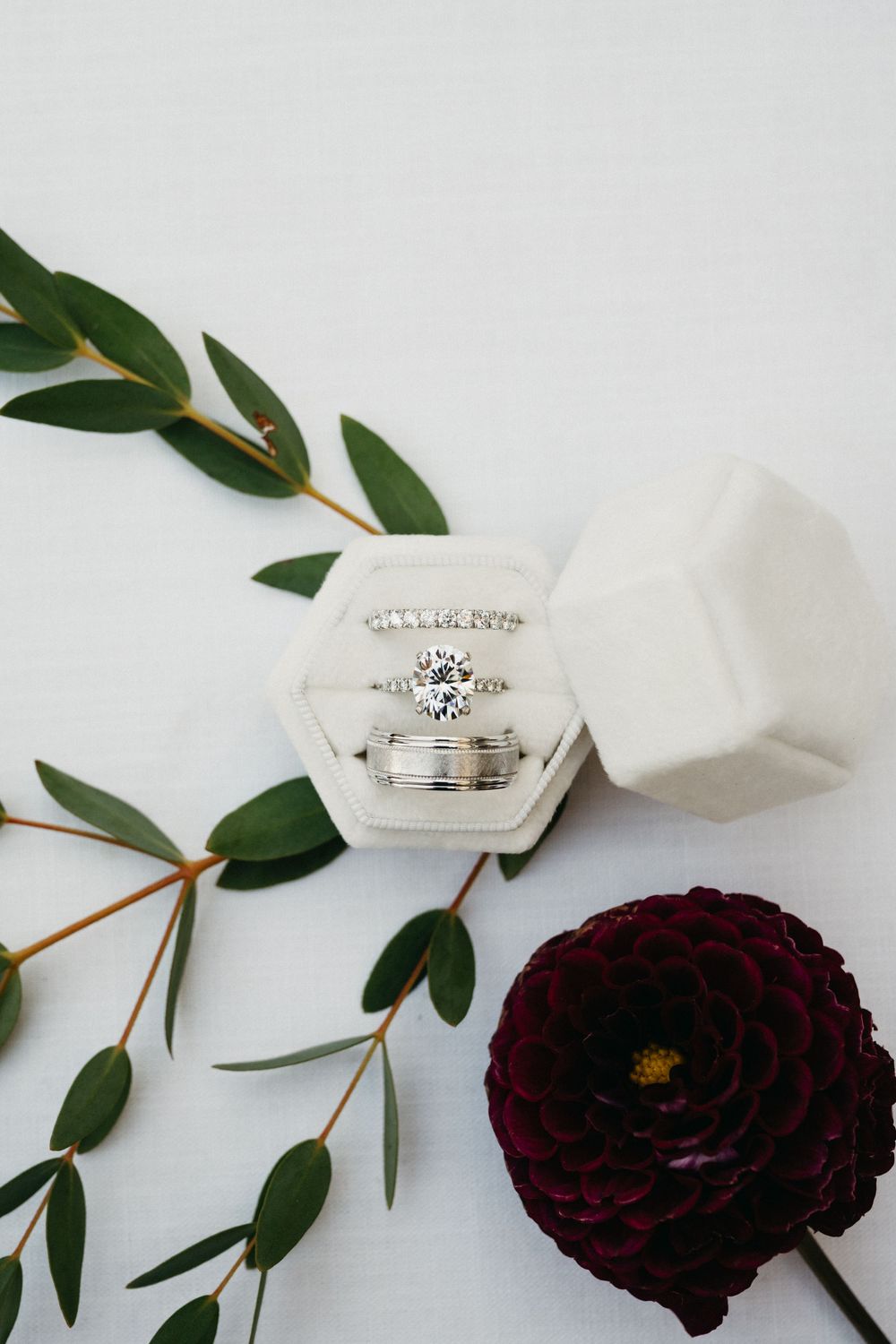 Wedding rings displayed in white hexagonal box alongside deep burgundy ranunculus flower and green foliage on white surface.