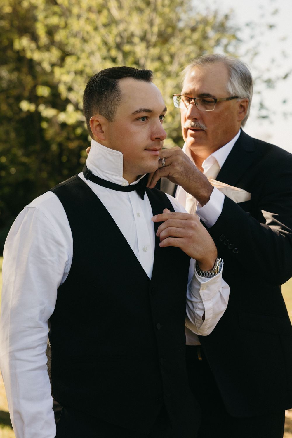 A tender moment of formal wear preparation outdoors as a bowtie is adjusted against a natural background.