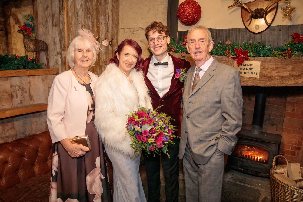 Wedding couple poses with family members in rustic indoor setting with fireplace and red balloon decor.