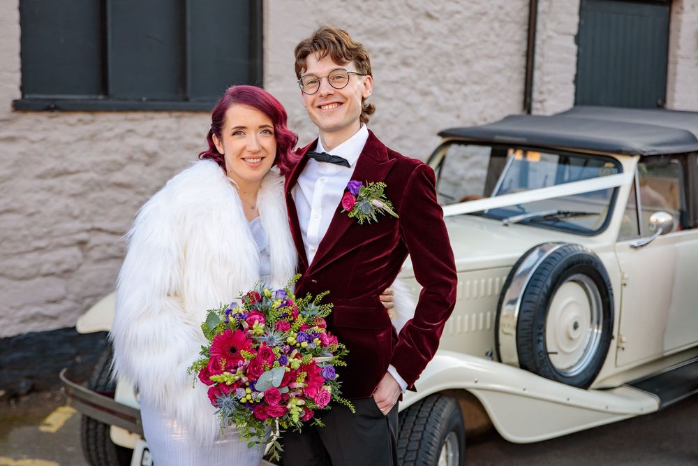 Couple in wedding attire poses next to vintage white car outdoors.