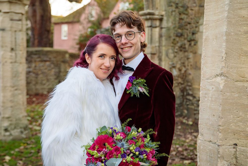 Wedding portrait under historic stone arch with vibrant bouquet of red and pink flowers.