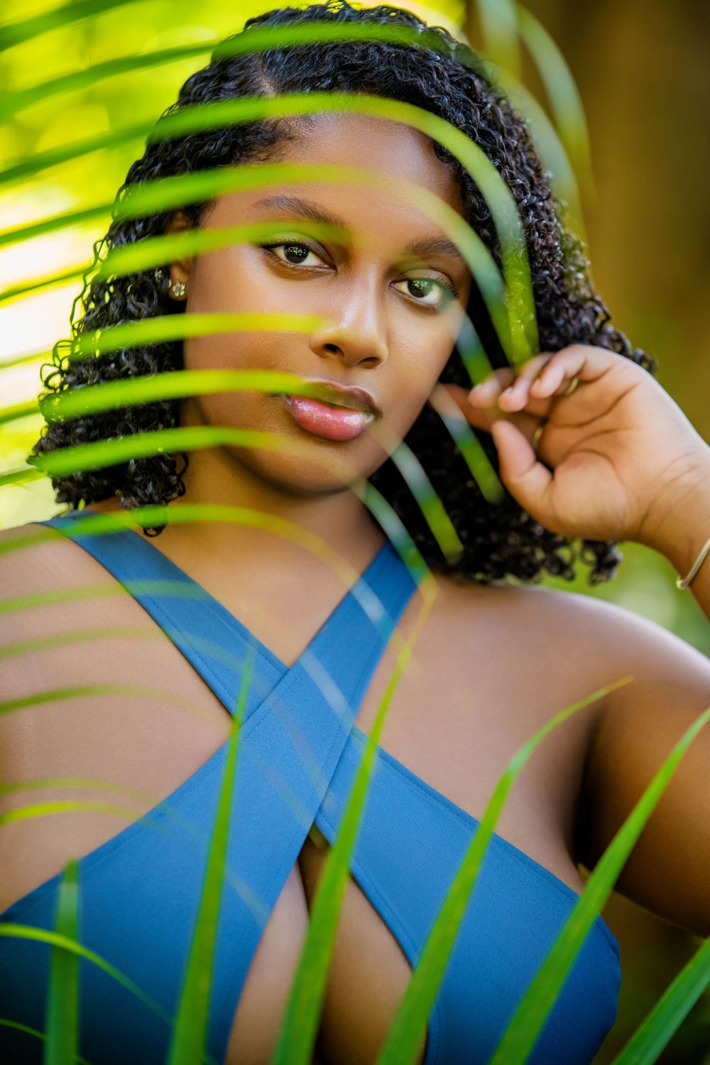 A portrait through palm leaves showing a person in a blue top with curly hair.