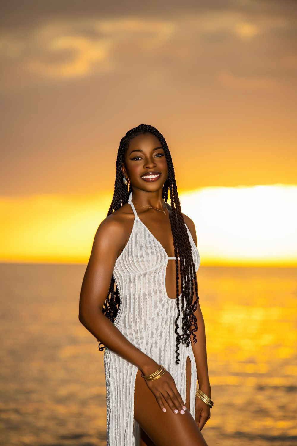 Model in white crochet dress poses against golden sunset over ocean.