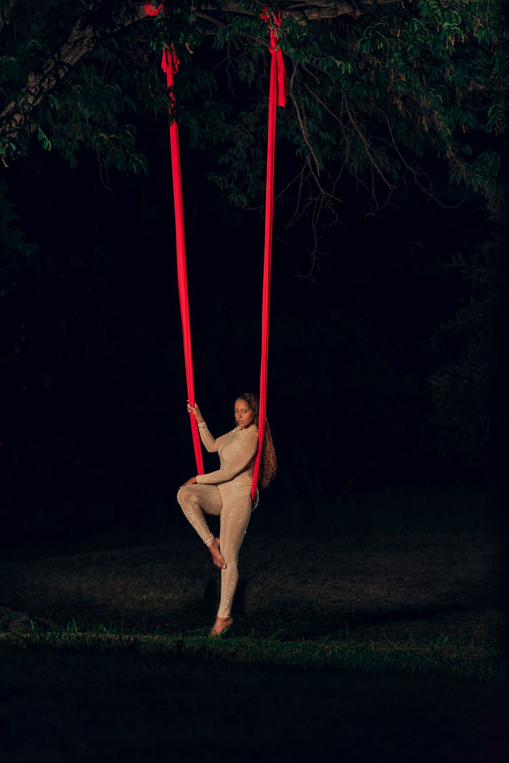 Performer gracefully poses on red aerial silks against dark outdoor backdrop.