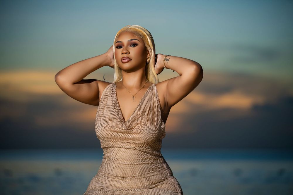 A person in a beige dress poses dramatically against a sunset sky backdrop.