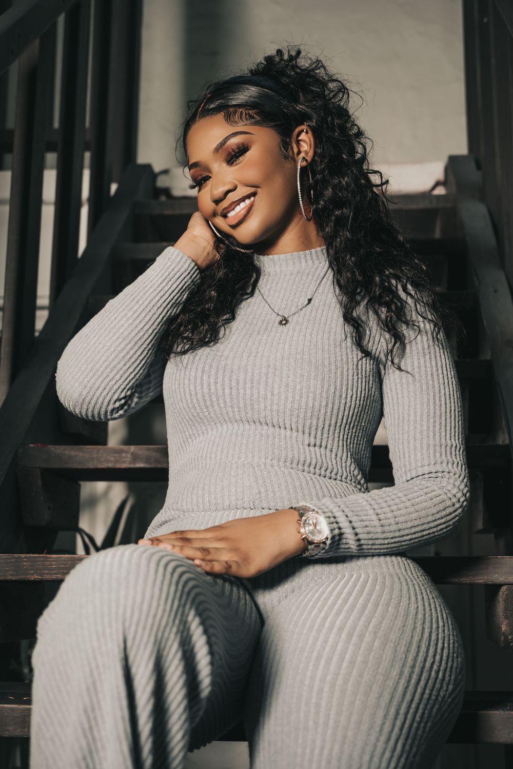 Model posing on stairs wearing grey ribbed loungewear set with long wavy hair.