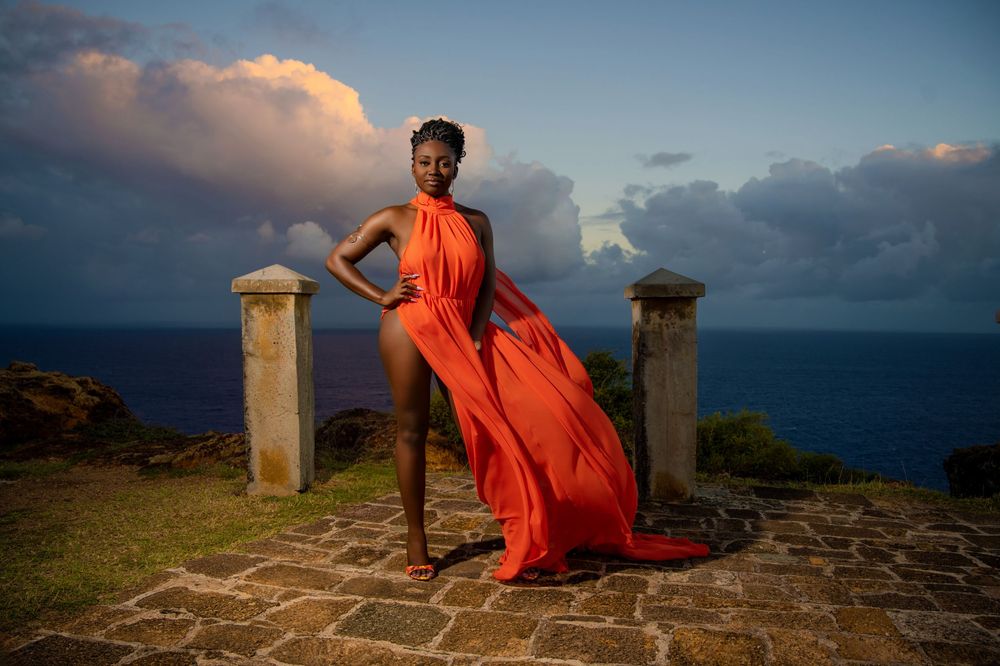 A person in a flowing orange dress poses dramatically against an ocean sunset backdrop.