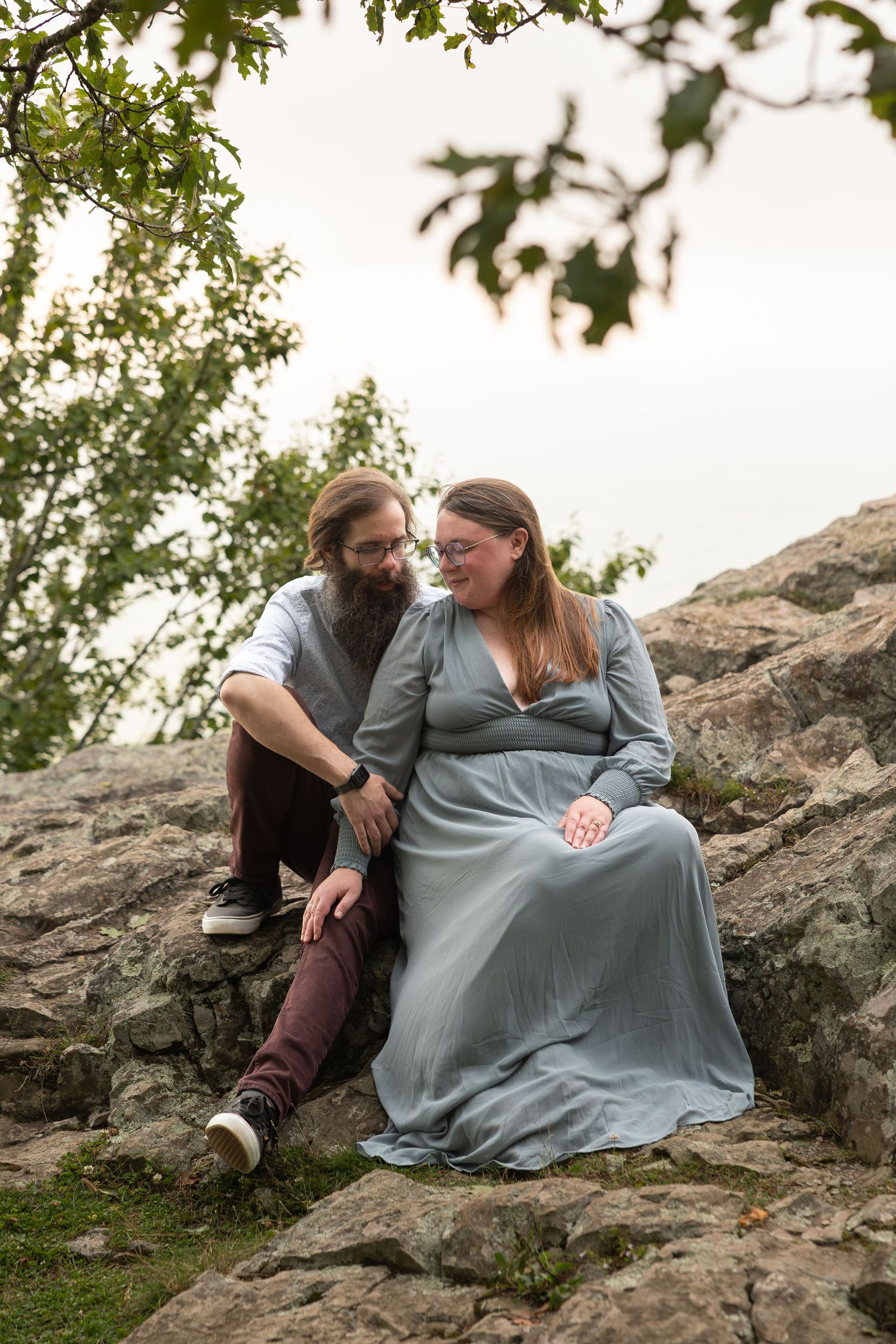 A couple sits together on a rocky mountain overlook during sunset as clouds gather in the dramatic sky.
