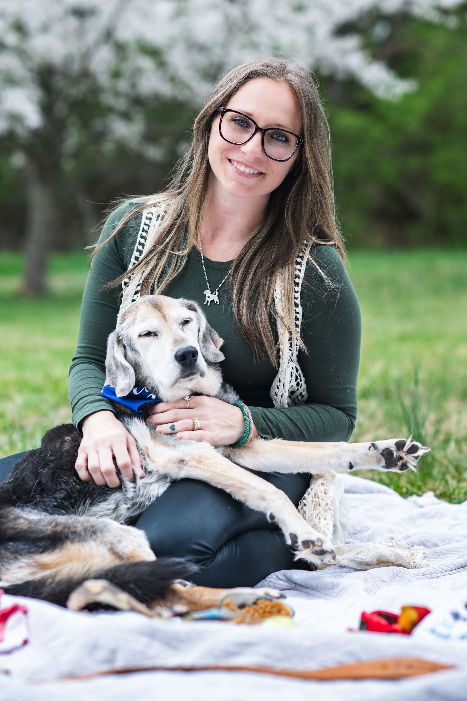 Senior dog with blue bow tie rests on blanket outdoors with person in green sweater during park picnic.