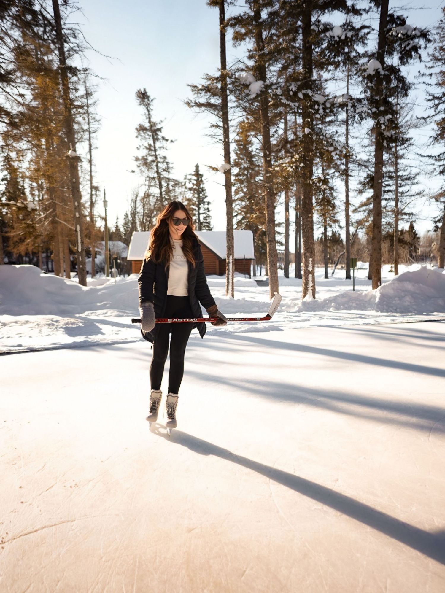 Person ice skating on a sunny winter path surrounded by tall pine trees.