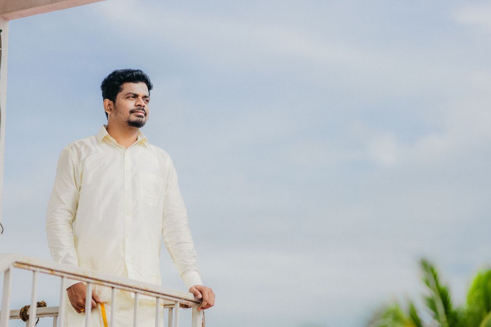 A person wearing a white shirt stands on a balcony against a blue cloudy sky with green plants visible.