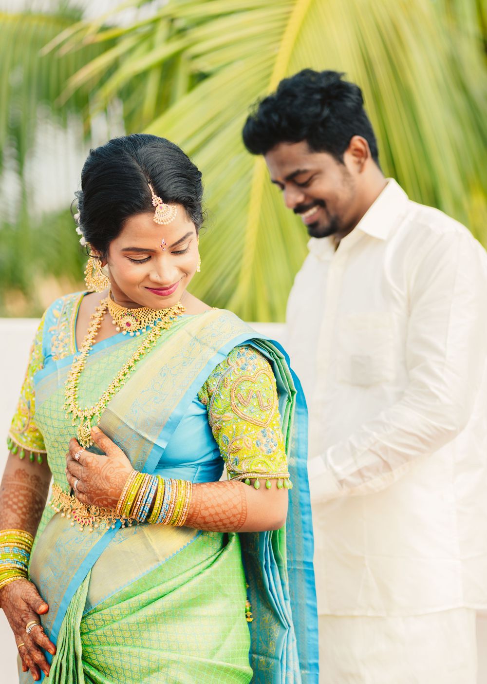 Traditional South Indian couple in wedding attire posing against palm leaves in bright natural lighting.