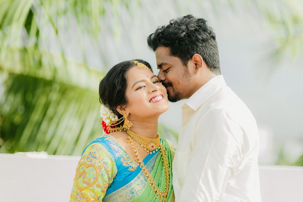 A couple in traditional Indian attire share an intimate moment against a white wall with green palm leaves in background.