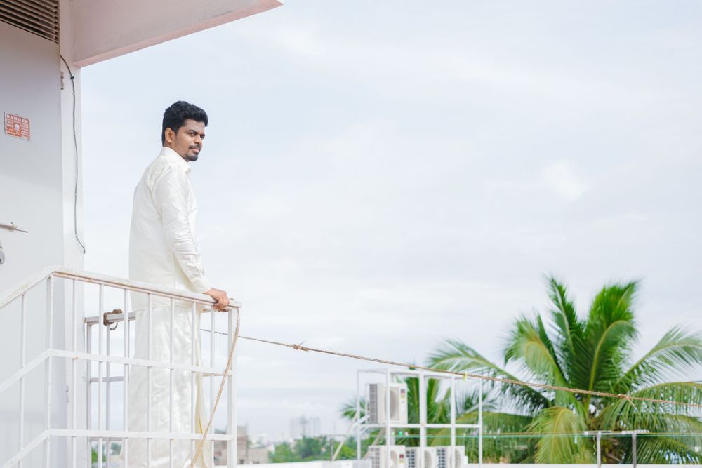 Man in white traditional attire standing on balcony with palm trees in background.